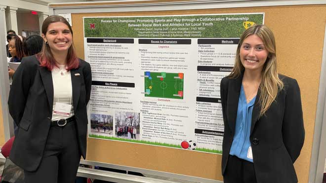 Two female students stand smiling in front of a large academic research poster titled “Recess for Champions,” which details a youth development program using sports and play. The poster includes sections on background, logistics, curriculum, and outcomes. The students are dressed in business attire and participating in Miami University’s Undergraduate Research Forum.