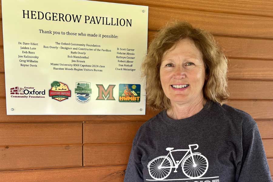 Miami University Professor Helaine Alessio stands beside a dedication plaque mounted on the pavilion wall at Hueston Woods State Park. The plaque lists donors and collaborators, including the Oxford Community Foundation, Hueston Woods Region Visitors Bureau, and Miami’s 2024 KNH capstone class, recognizing their role in creating the new community pavilion.