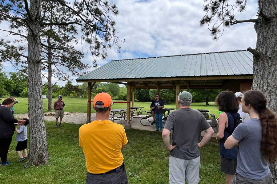 A small group of people stands in front of a new wooden pavilion at Hueston Woods State Park during a ribbon-cutting event. Miami University Professor Helaine Alessio addresses the crowd, celebrating the collaborative completion of the pavilion by her KNH capstone students, community members, and volunteers, supported by a grant from the Oxford Community Foundation.