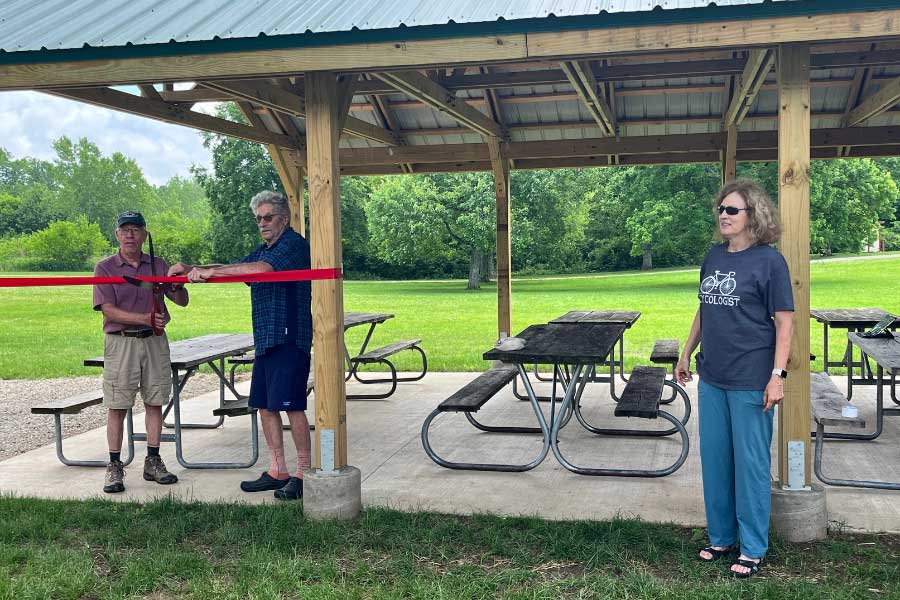 Two community members hold a red ribbon in front of the new pavilion at Hueston Woods State Park, preparing for the ceremonial ribbon-cutting. Miami University Professor Helaine Alessio stands to the right, smiling, as the group marks the official opening of the space built with support from her capstone students and local partners.