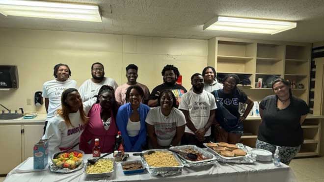Thirteen TEACh participants posing behind a table filled with food dishes and drinks in a community kitchen.