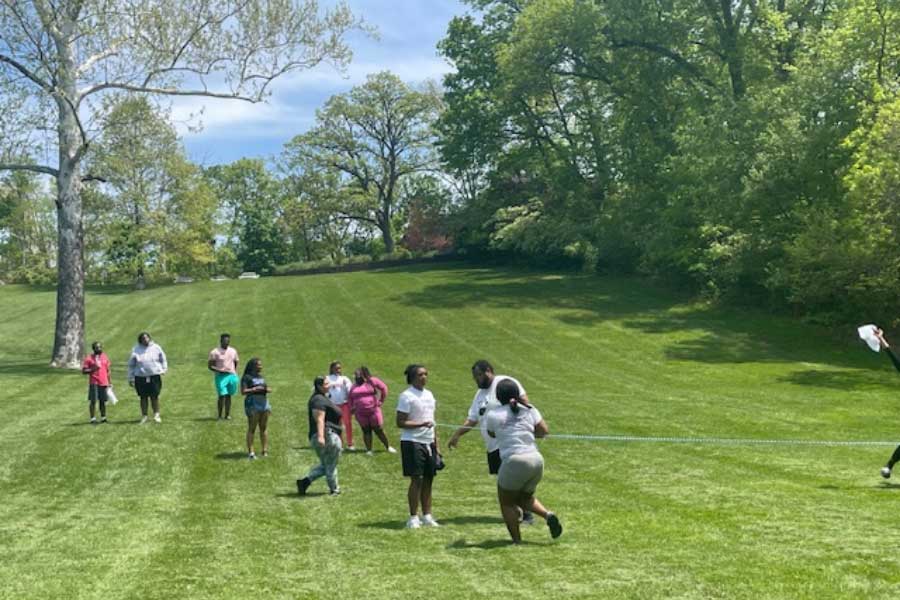 A group of people spread out across a large grassy field on a sunny day, with some in the foreground playing or participating in an activity involving a rope or line on the ground.