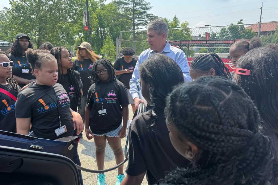 Kevin Kushman, Joyce Barnes Farmer Guest Professor at Miami University, speaks to a group of middle schoolers from Cincinnati Public Schools during Camp Excel. The students, wearing matching black program shirts, surround Kushman as he discusses electric vehicle technology in an outdoor campus parking area.
