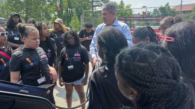 Kevin Kushman, Joyce Barnes Farmer Guest Professor at Miami University, speaks to a group of middle schoolers from Cincinnati Public Schools during Camp Excel. The students, wearing matching black program shirts, surround Kushman as he discusses electric vehicle technology in an outdoor campus parking area.