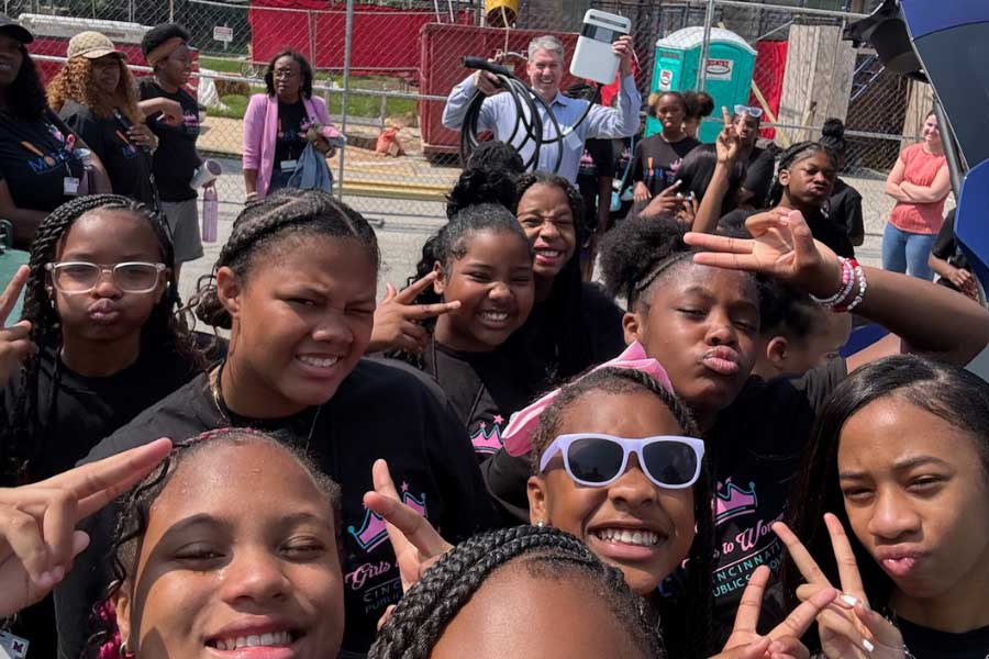 A cheerful group selfie of middle school girls from Cincinnati Public Schools attending Camp Excel. The students, wearing matching black shirts, smile, flash peace signs, and enjoy the sunny day at Miami University. Kevin Kushman appears in the background holding an EV charger cable, smiling as the group poses.