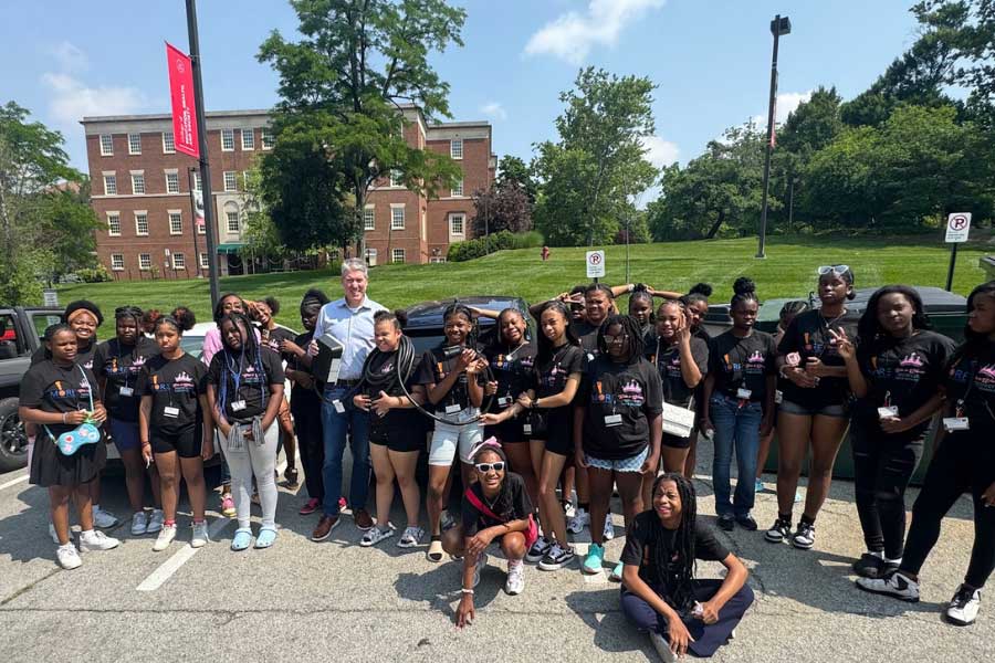 Kevin Kushman stands with a large group of Camp Excel participants—middle schoolers from Cincinnati Public Schools—outside on Miami University’s campus. The students, dressed in black program t-shirts, gather around him smiling for a group photo with a red university banner and academic building in the background.