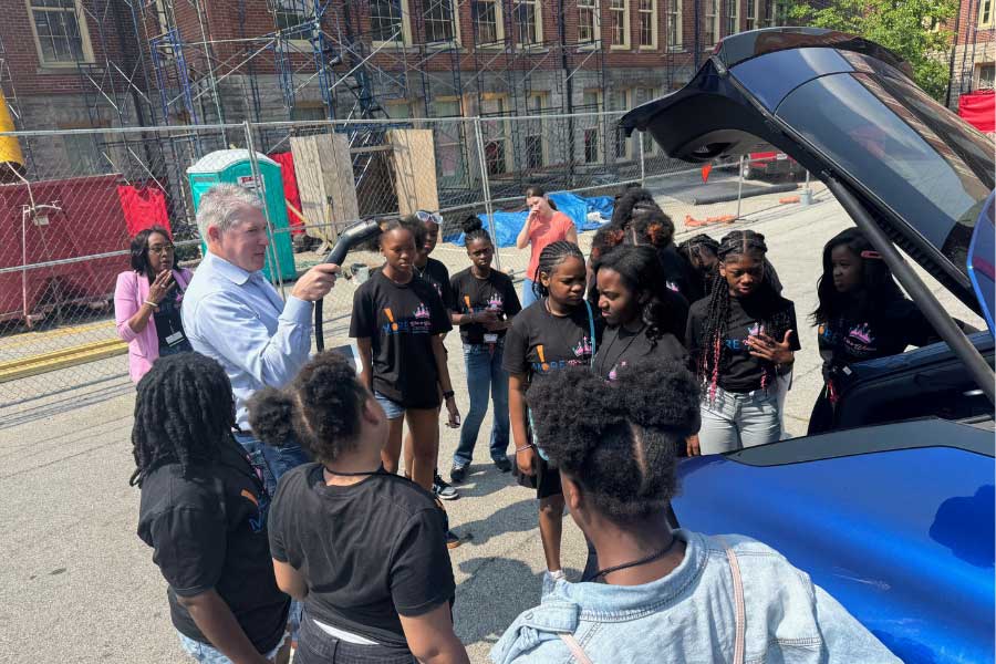 Kevin Kushman demonstrates electric vehicle technology to Camp Excel students during their visit to Miami University. Standing next to an EV with the trunk open, Kushman explains a component while students from Cincinnati Public Schools listen attentively, gathered in a semicircle on a sunny campus day.