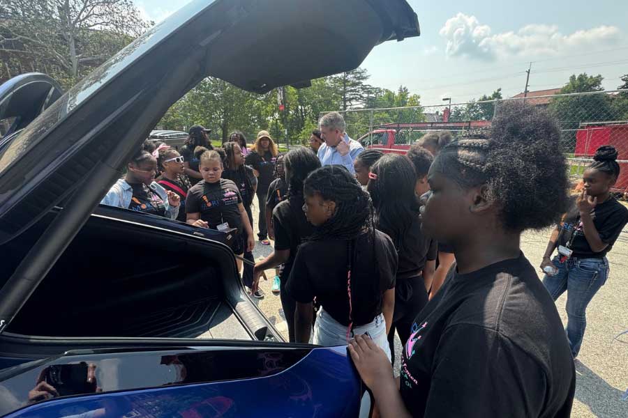 Kushman discusses electric vehicles beside a blue EV with the trunk open. Kushman gestures while addressing the group, sharing insights from his journey in innovation and entrepreneurship.