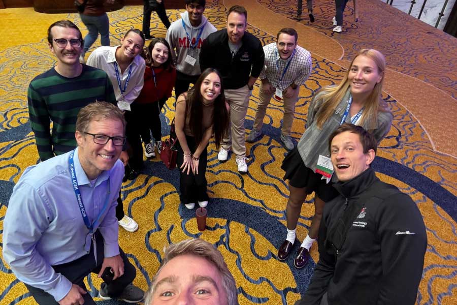 Group of Miami University students and faculty smiling at a conference, gathered on a patterned carpet.