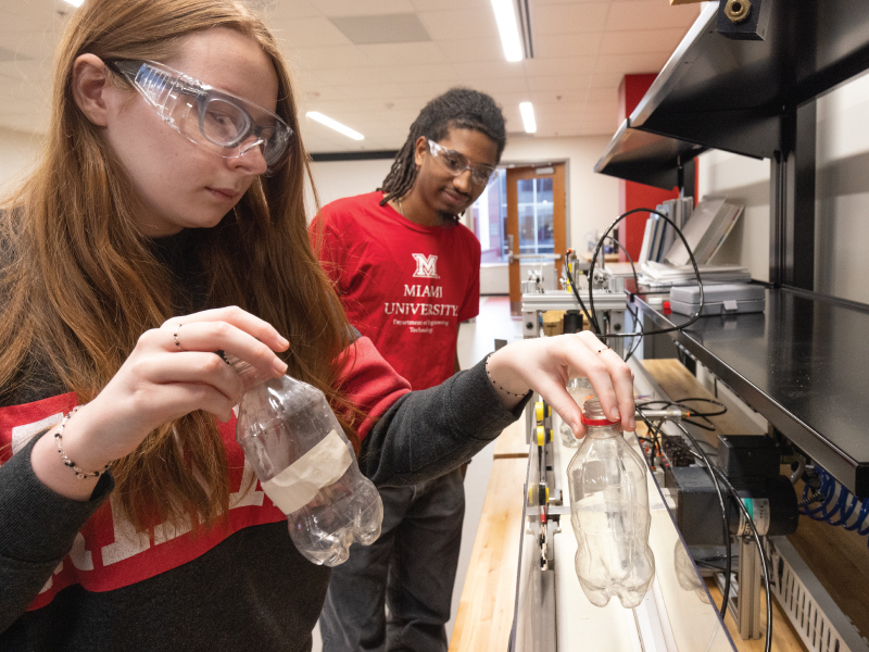 2 students using a empty bottle and a piece of equipment at the Advanced Manufacturing Workforce and Innovation Hub.