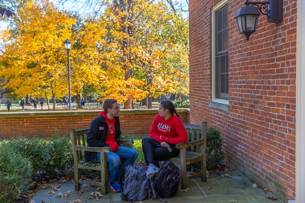 Photo of two student sitting outside of a residence hall with their backpacks at their feet.