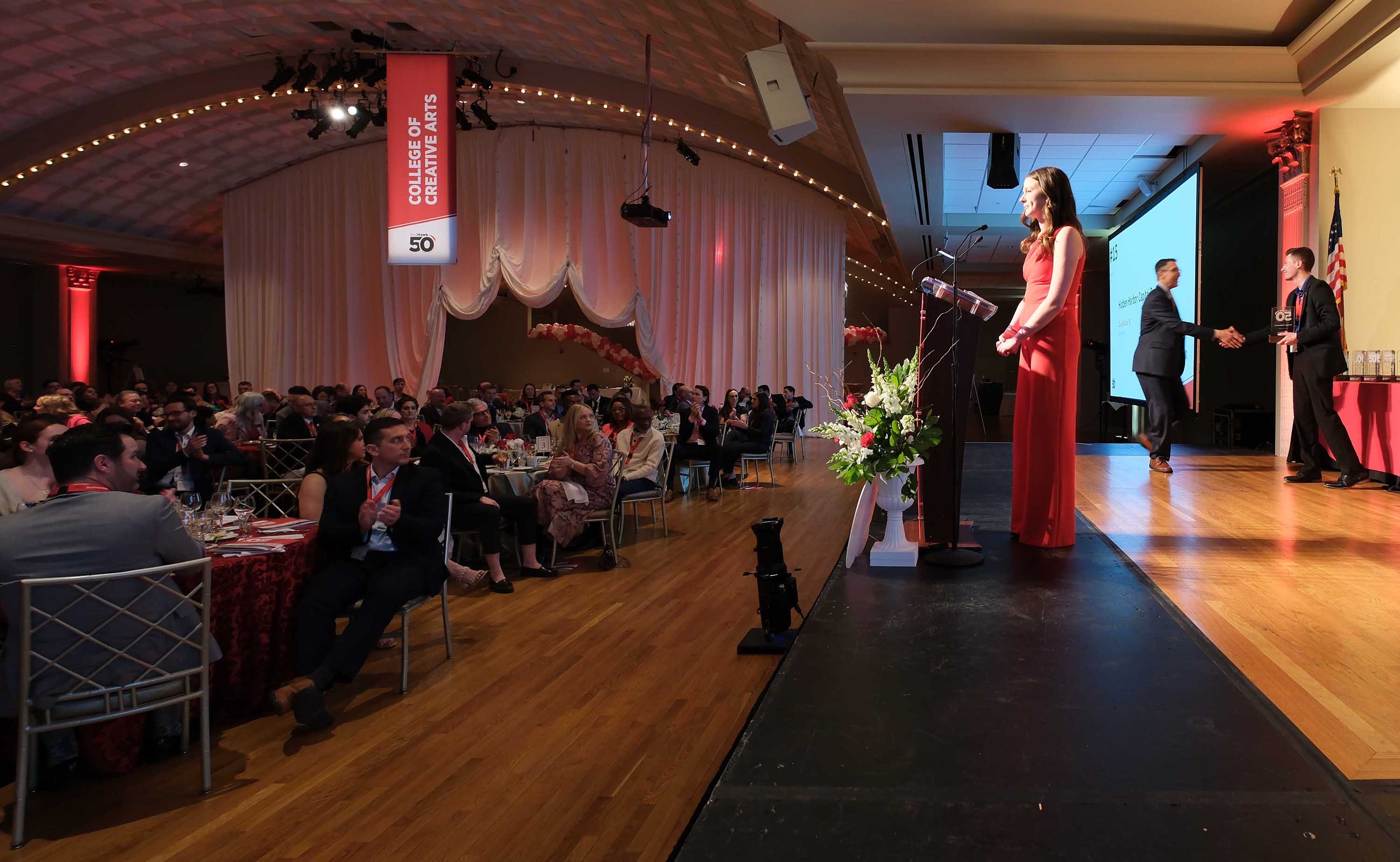 Wide shot of presentation, crowd to left, honoree on stage to right