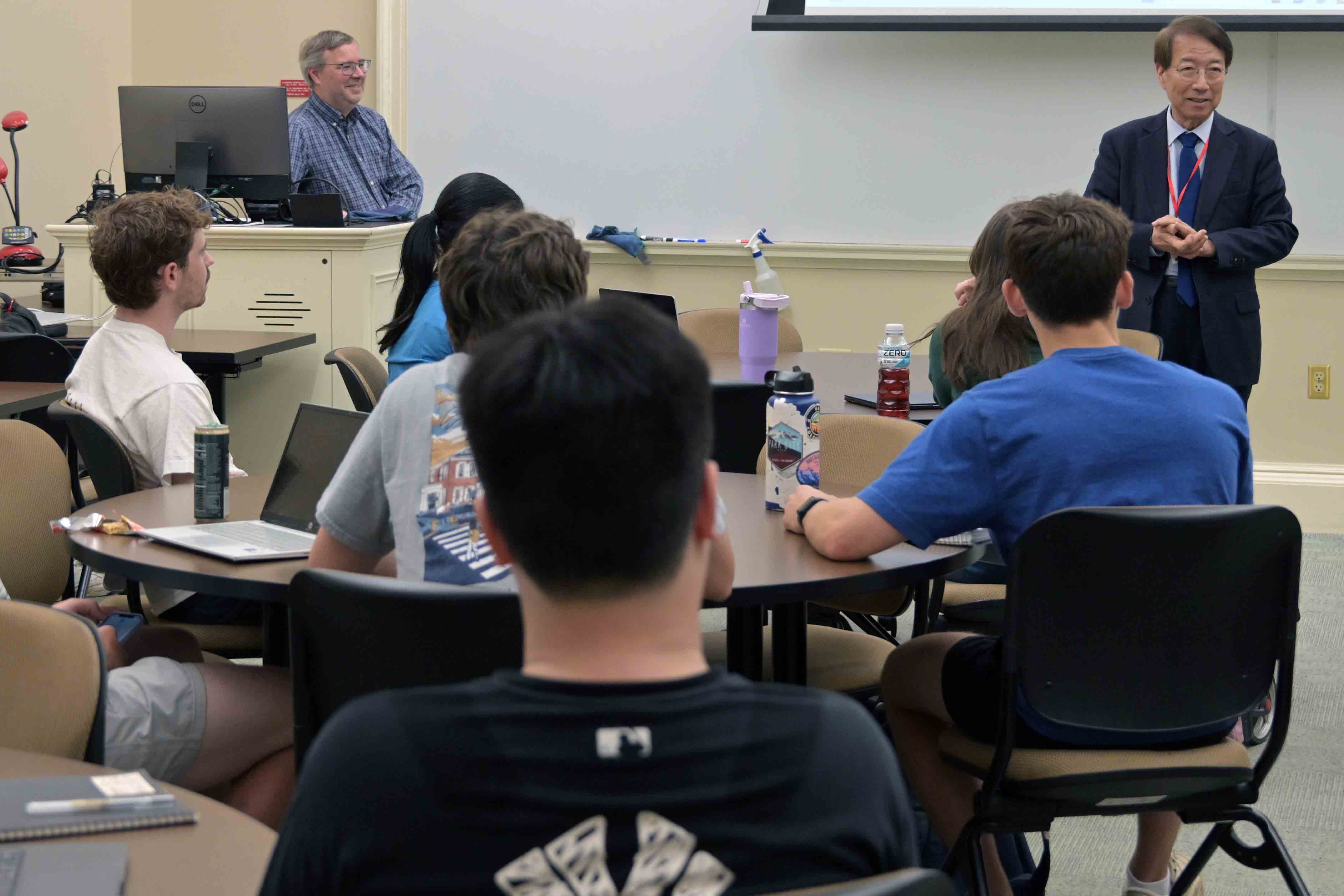 A Higgin Kim presenter gives a lecture to a group of students seated at tables while another instructor observes from the back of the classroom.