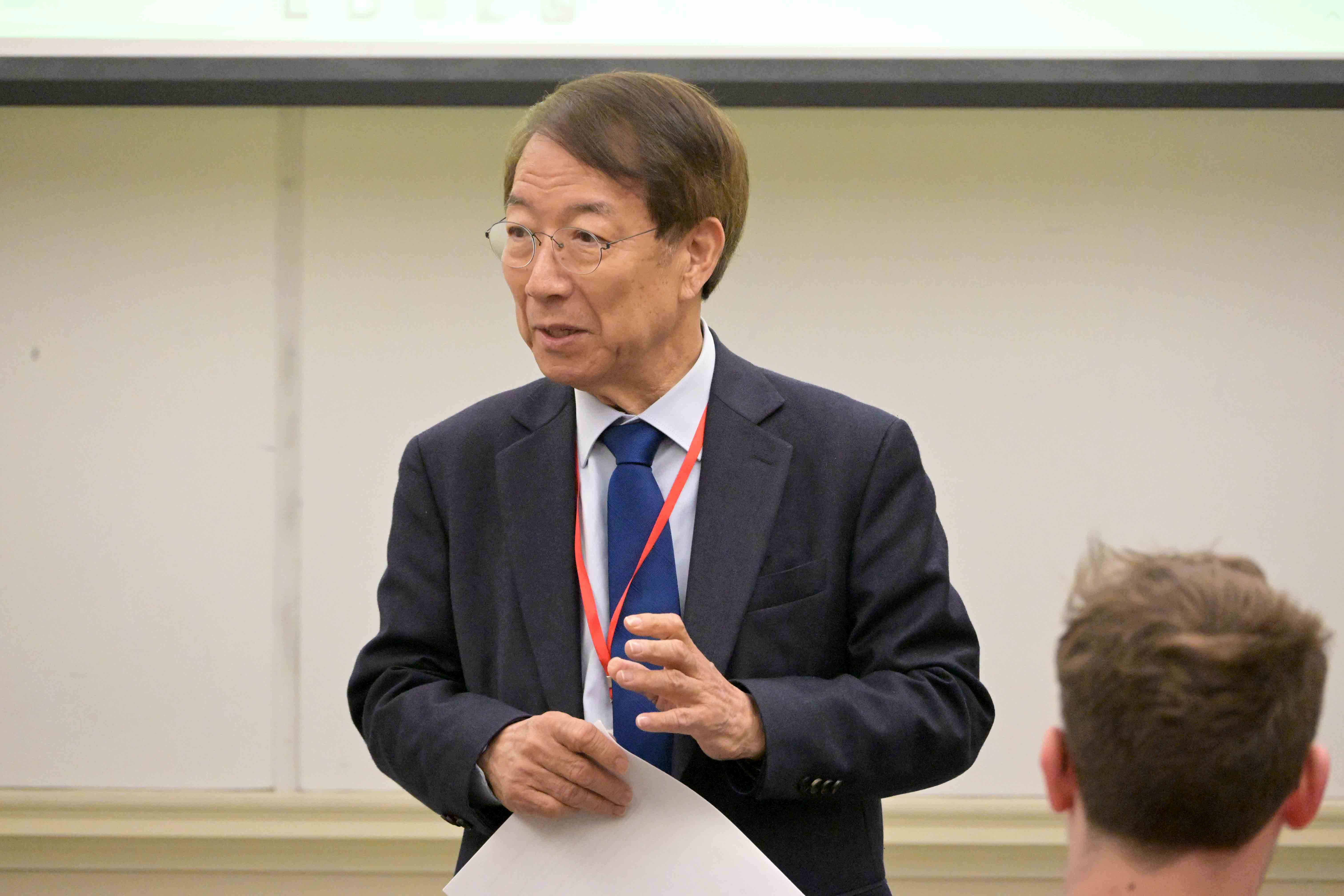 A speaker presenting at a conference, wearing glasses and a suit, with a name badge visible around his neck.