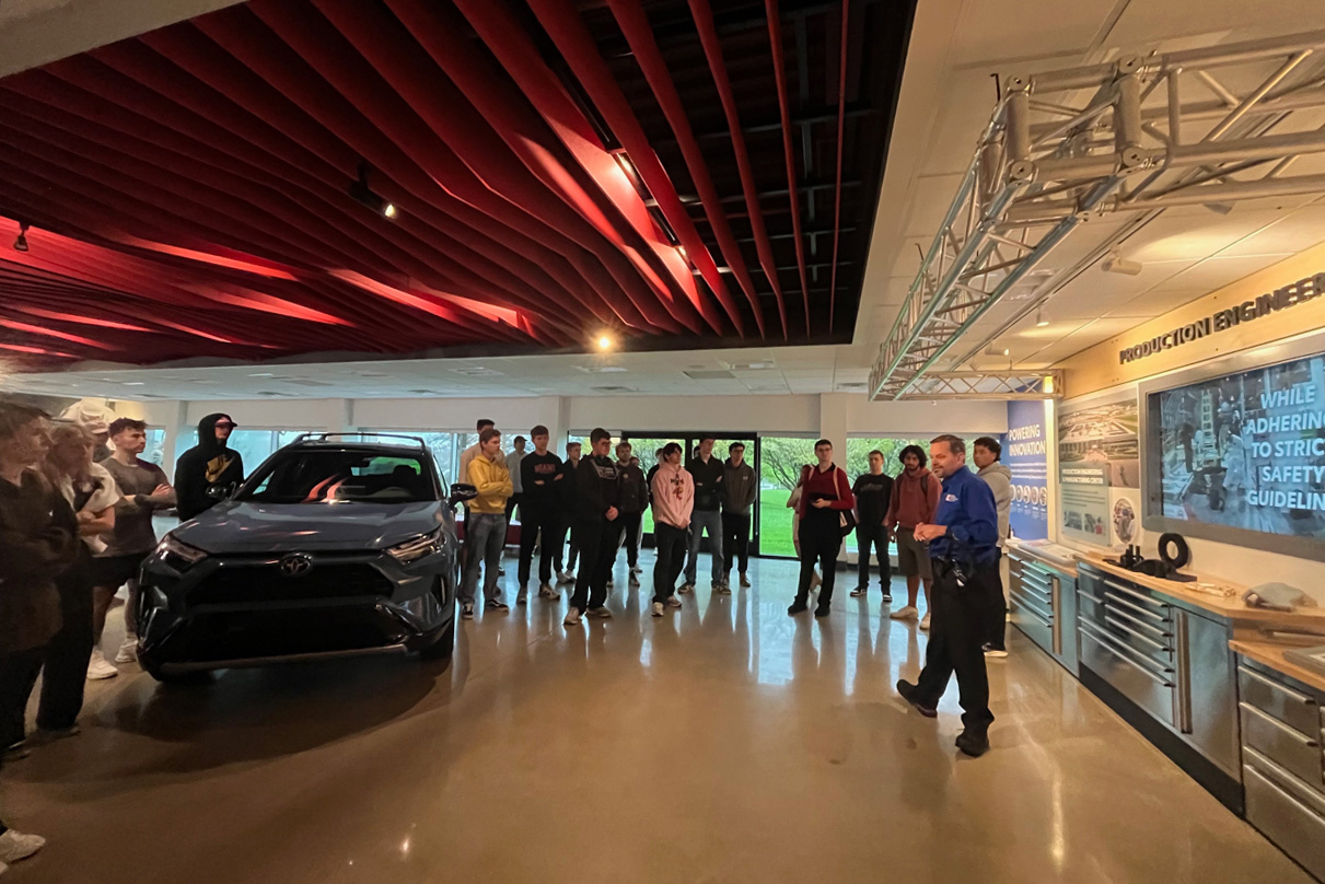 A group of students attentively listens to a presenter in an automotive engineering facility, with a vehicle displayed in the foreground.