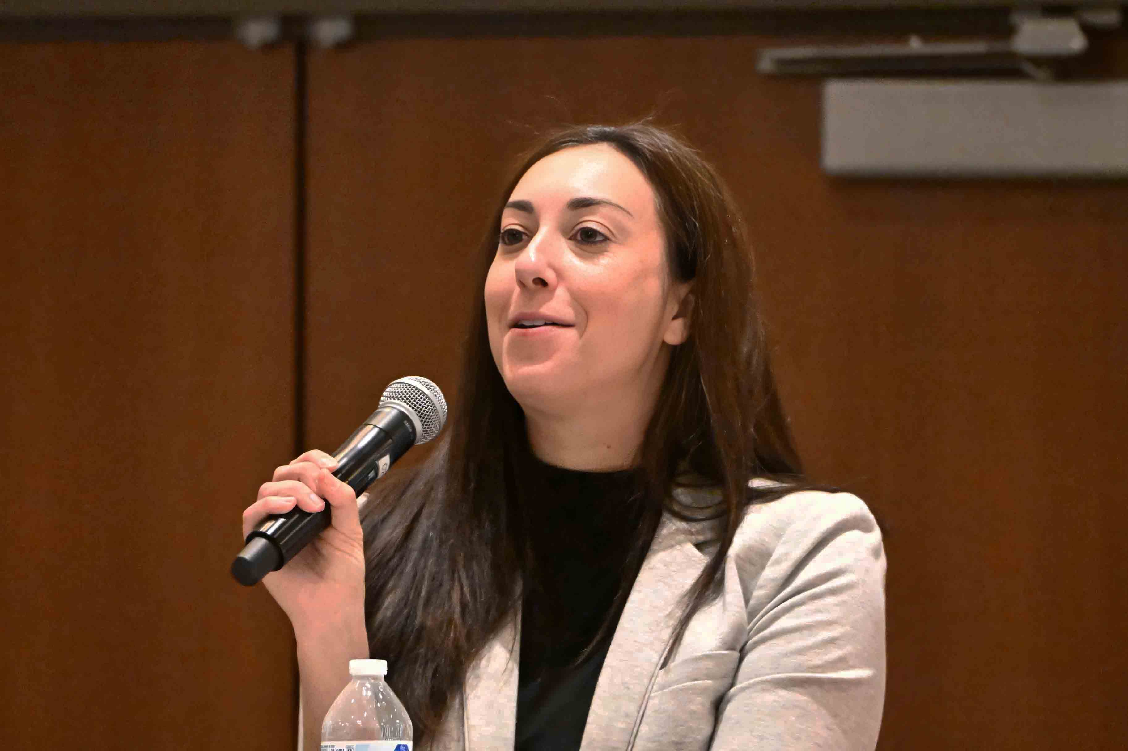 Jessica Bhadauria speaking into a microphone at a panel discussion, dressed in a light gray blazer.