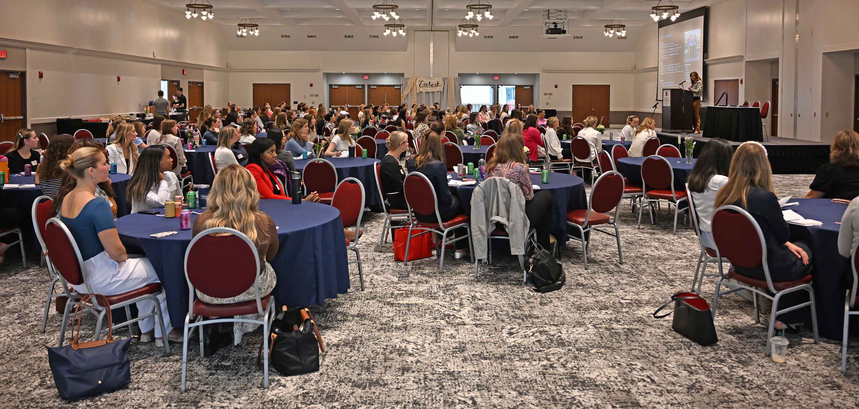 A large audience of attendees seated at round tables during the conference as Abell speaks