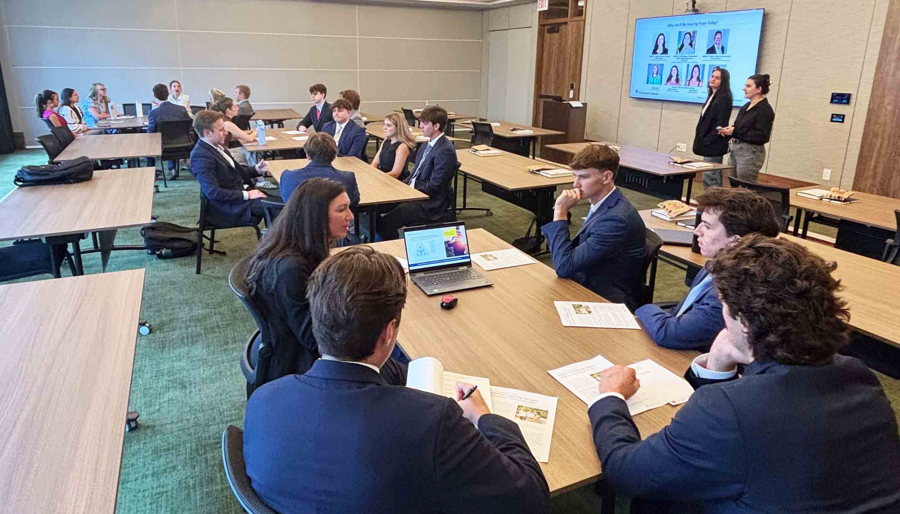 Group of students engaged in a discussion during a classroom presentation, with a teacher addressing the class in a modern conference room.