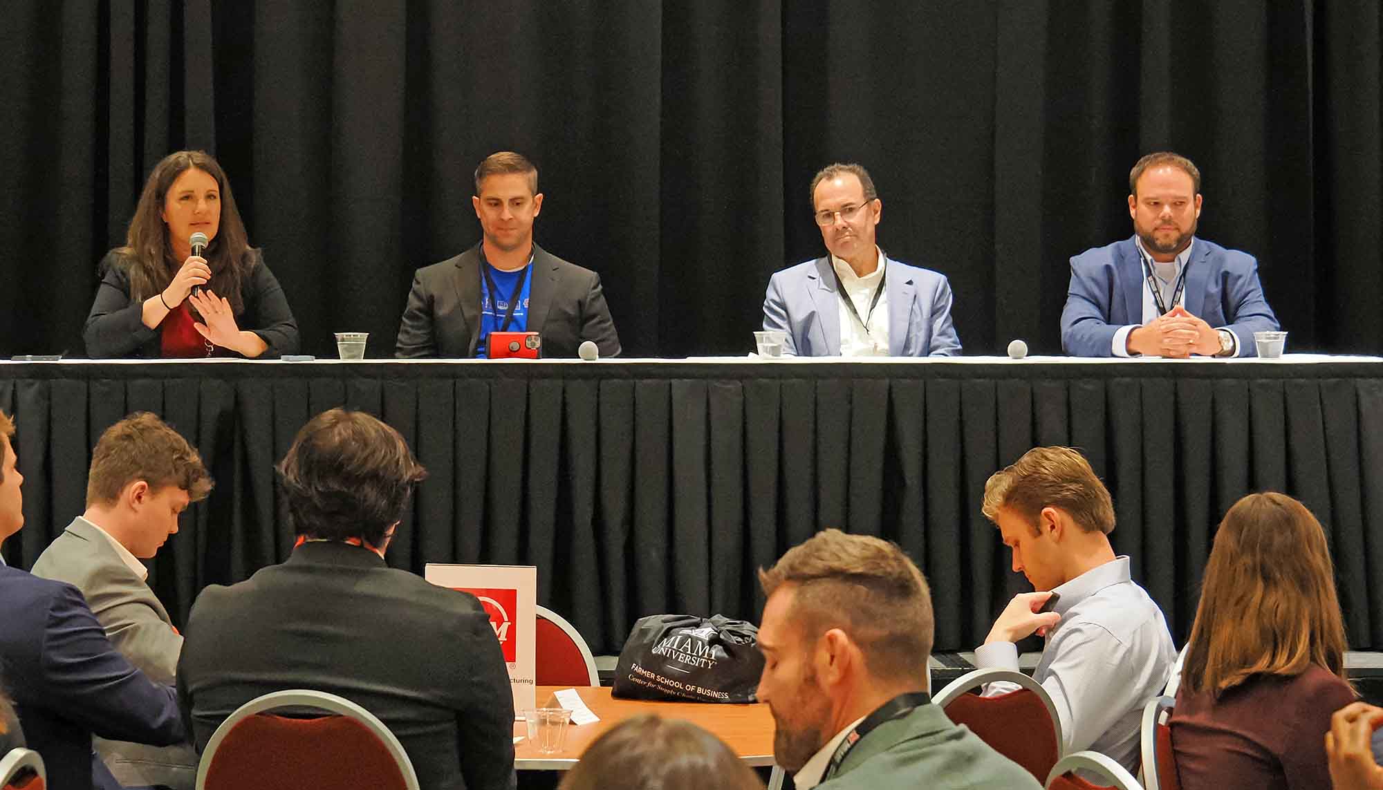 Panel discussion featuring four speakers at a conference, with one woman speaking into a microphone and three men seated beside her, while the audience is attentively listening.