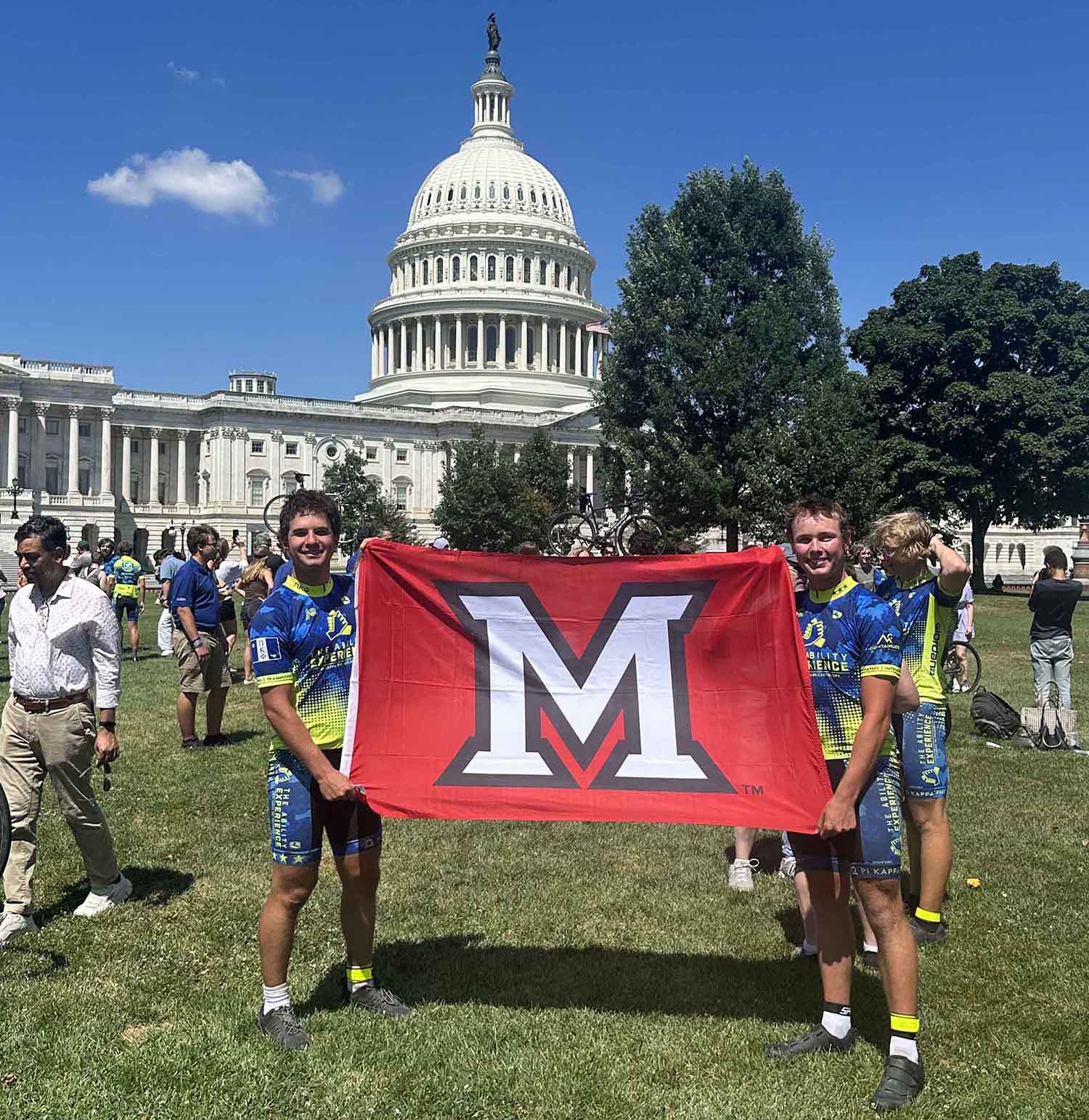 Two cyclists holding a red flag with an "M" logo in front of the U.S. Capitol building.
