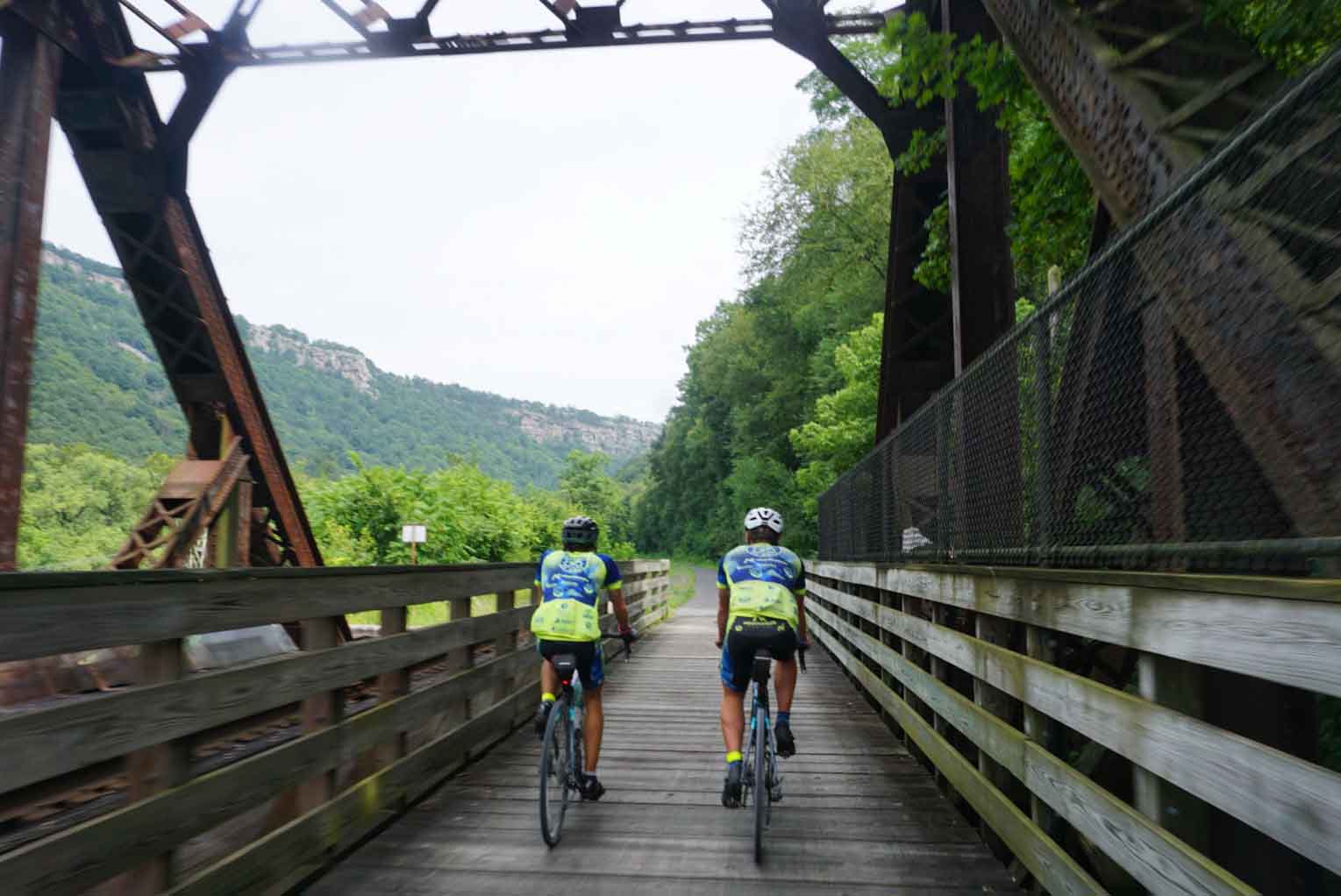 Two cyclists ride on a wooden bridge surrounded by greenery, with hills visible in the background.