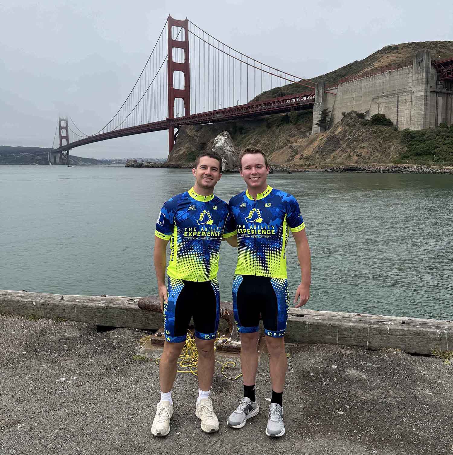 Two cyclists in vibrant blue and yellow jerseys stand by the waterfront, with the Golden Gate Bridge in the background.