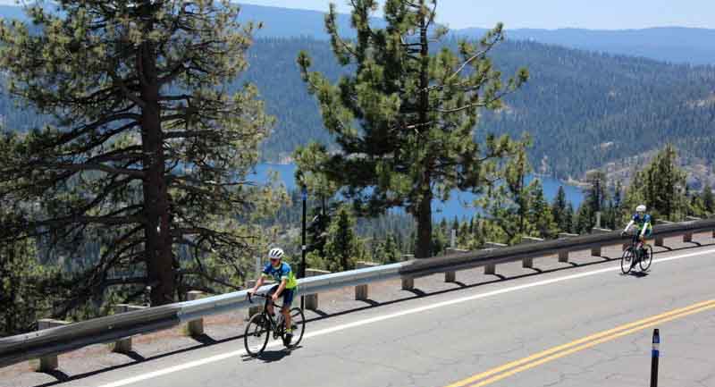 Two cyclists riding along a mountain road with a lake and pine trees in the background.