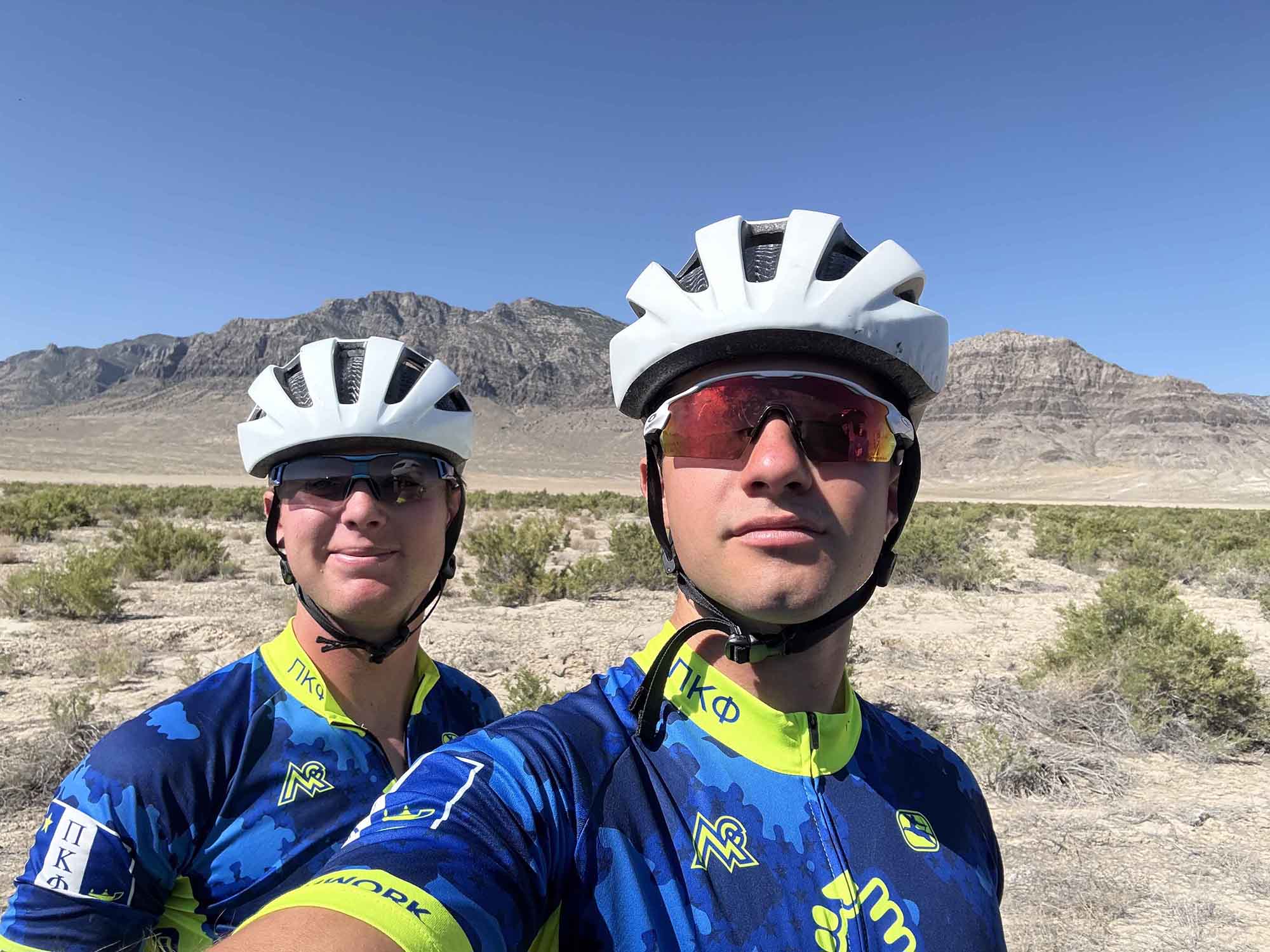 Two cyclists posing for a selfie in a desert landscape with mountains in the background. They are wearing matching blue and green cycling jerseys and helmets.