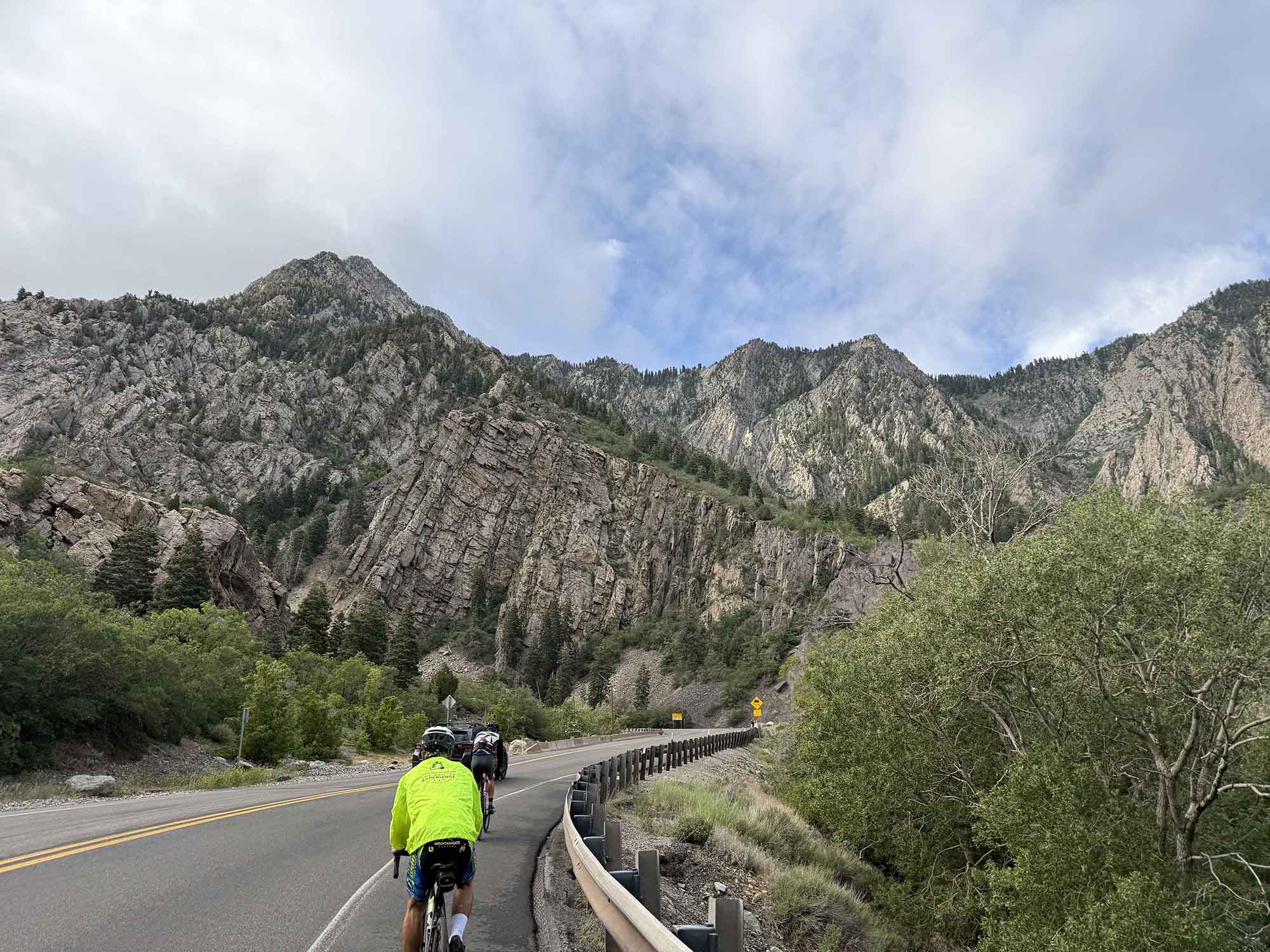 A group of cyclists rides along a winding road with dramatic mountain scenery in the background.