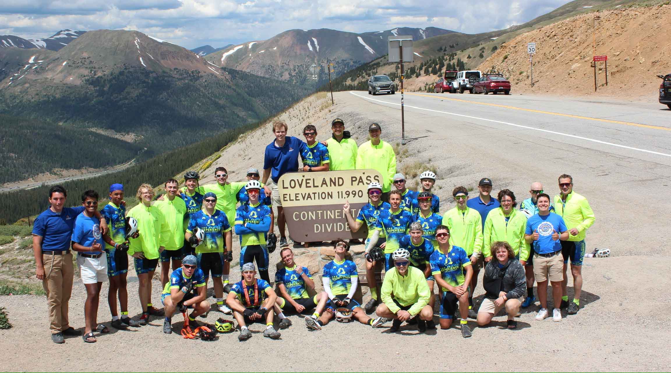 A large group of cyclists in bright jerseys and helmets poses at Loveland Pass, showcasing the elevation sign and scenic mountain backdrop.