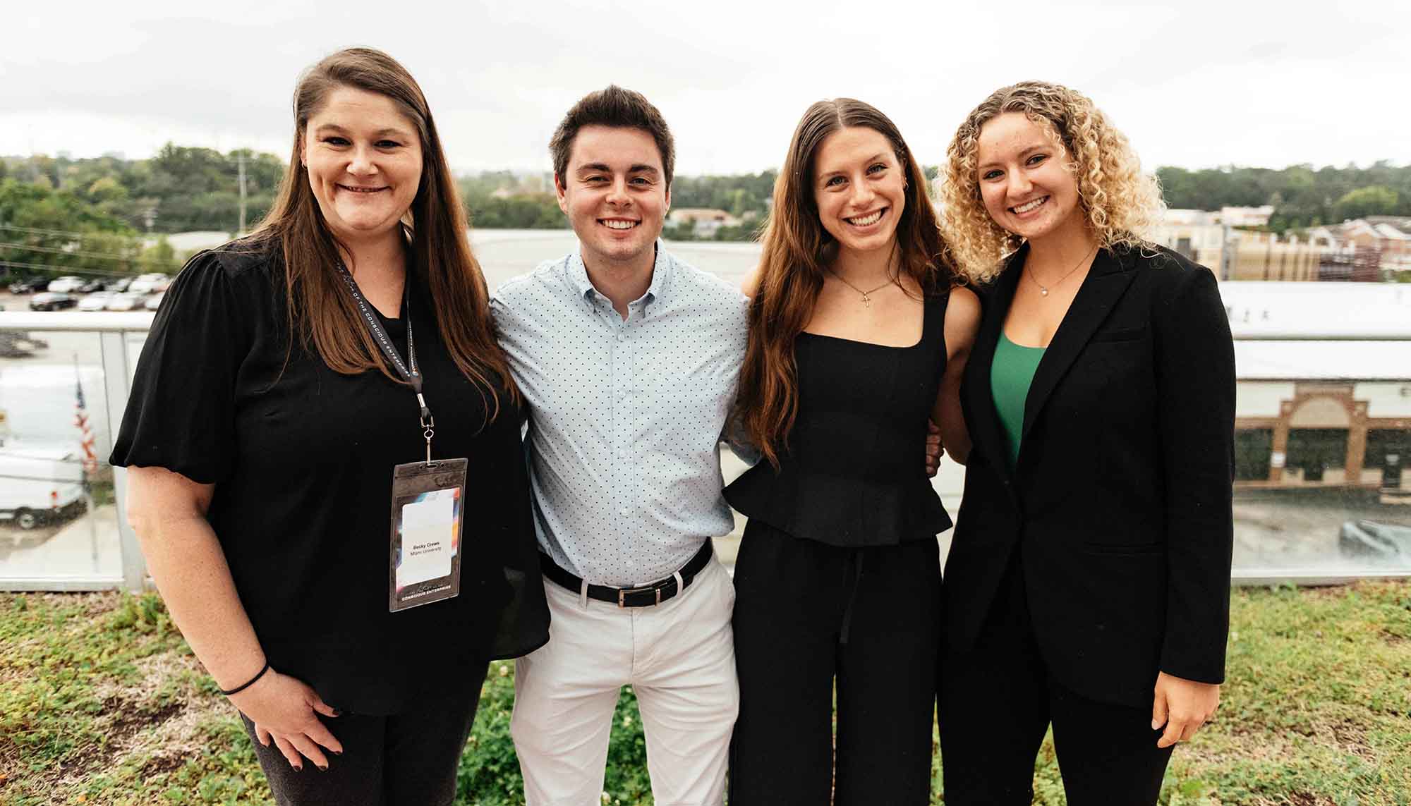 A group of four young adults smiling together outdoors, dressed in professional attire, with a scenic background.