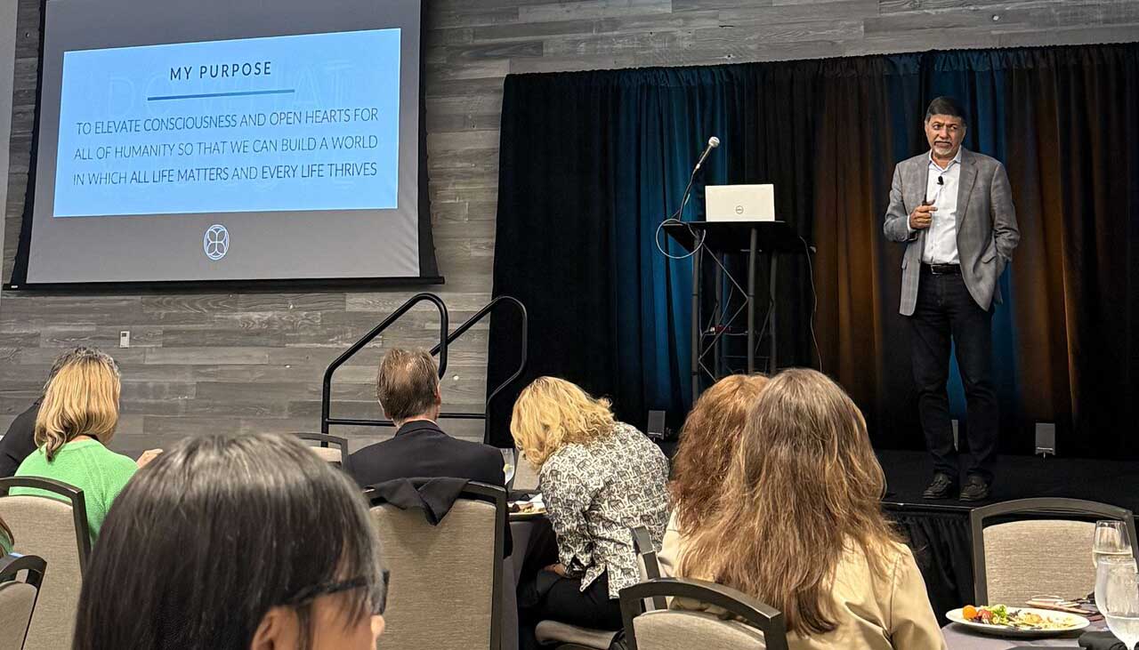 A speaker presenting on stage with an audience, sharing a message about elevating consciousness and opening hearts for humanity.