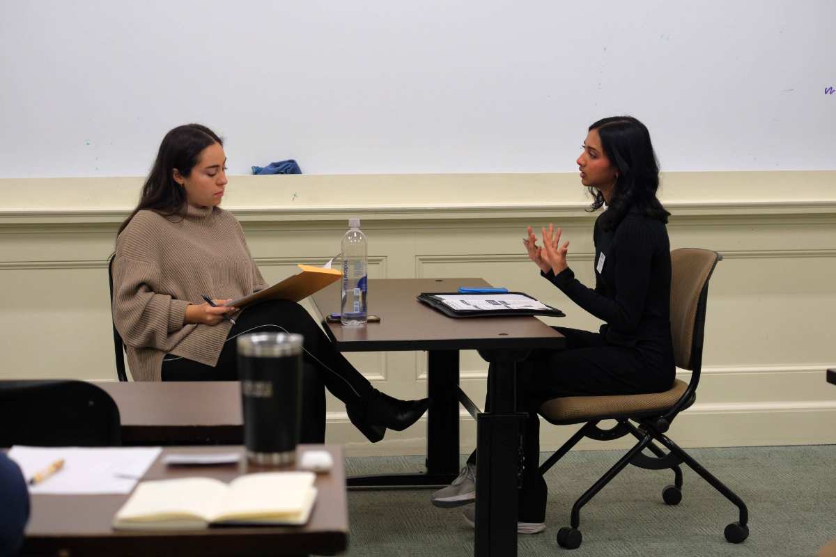 Two young women engaged in a conversation during a classroom discussion or interview setting. One is holding a folder while the other is speaking intently.