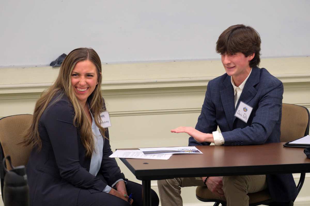 A young man and a woman sit at a table engaged in a conversation, both smiling and wearing professional attire.