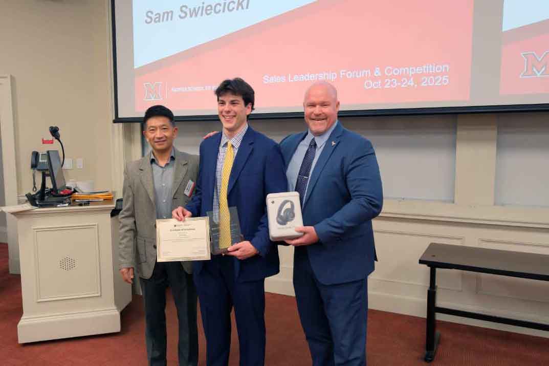Three individuals pose together at a Sales Leadership Forum, with one holding a certificate and another holding a gift bag with headphones.