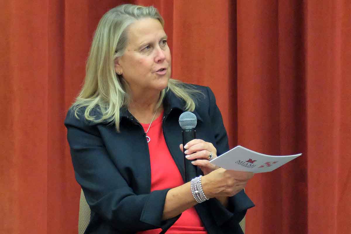 A woman in a black blazer speaks into a microphone while holding a program, with a red curtain in the background.