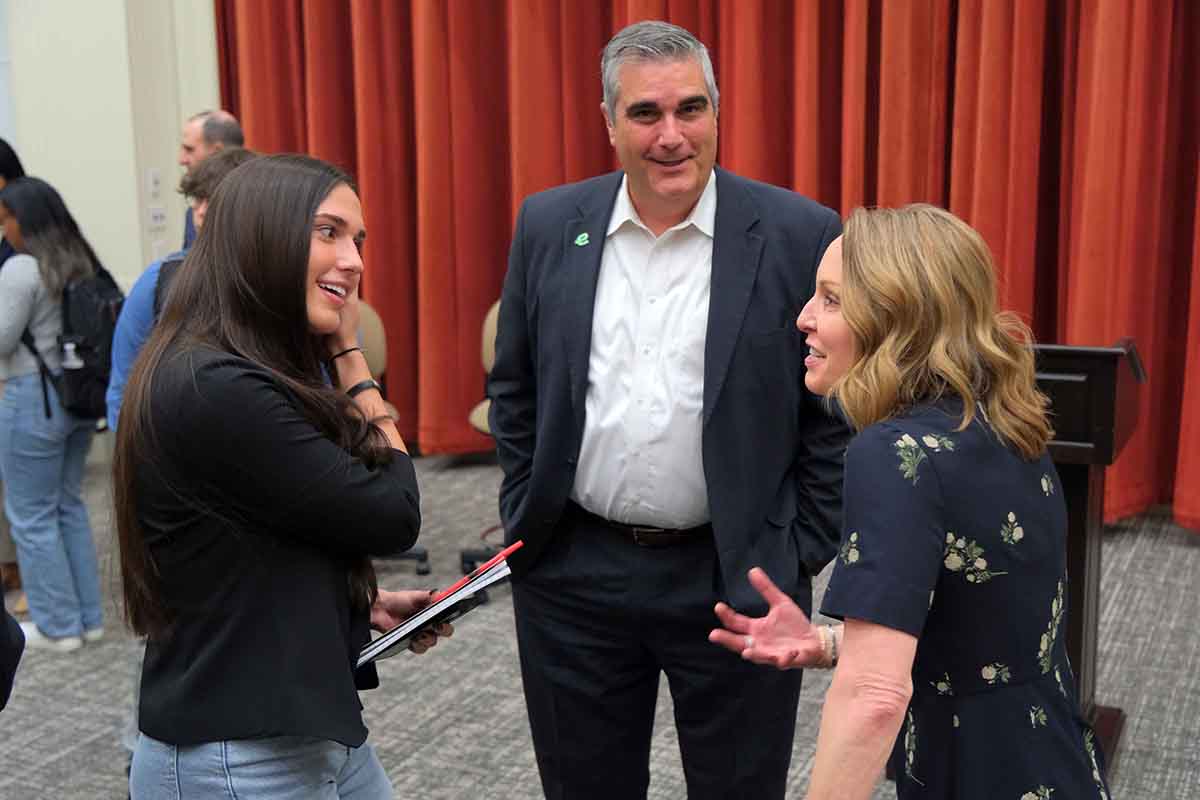 Three individuals engaged in conversation at a networking event, with two women smiling and a man listening attentively.