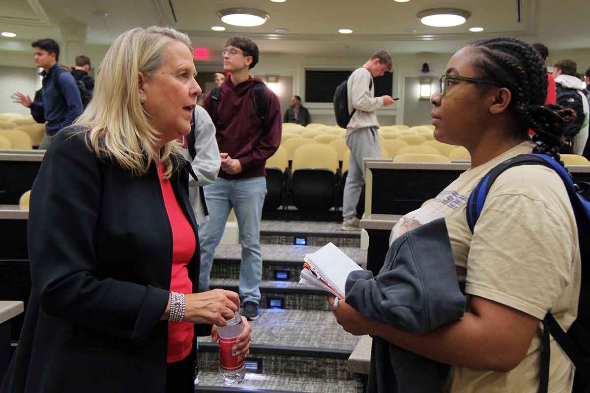 A woman in a black blazer engages in conversation with a student in a beige t-shirt, set in a classroom environment with empty seats in the background.