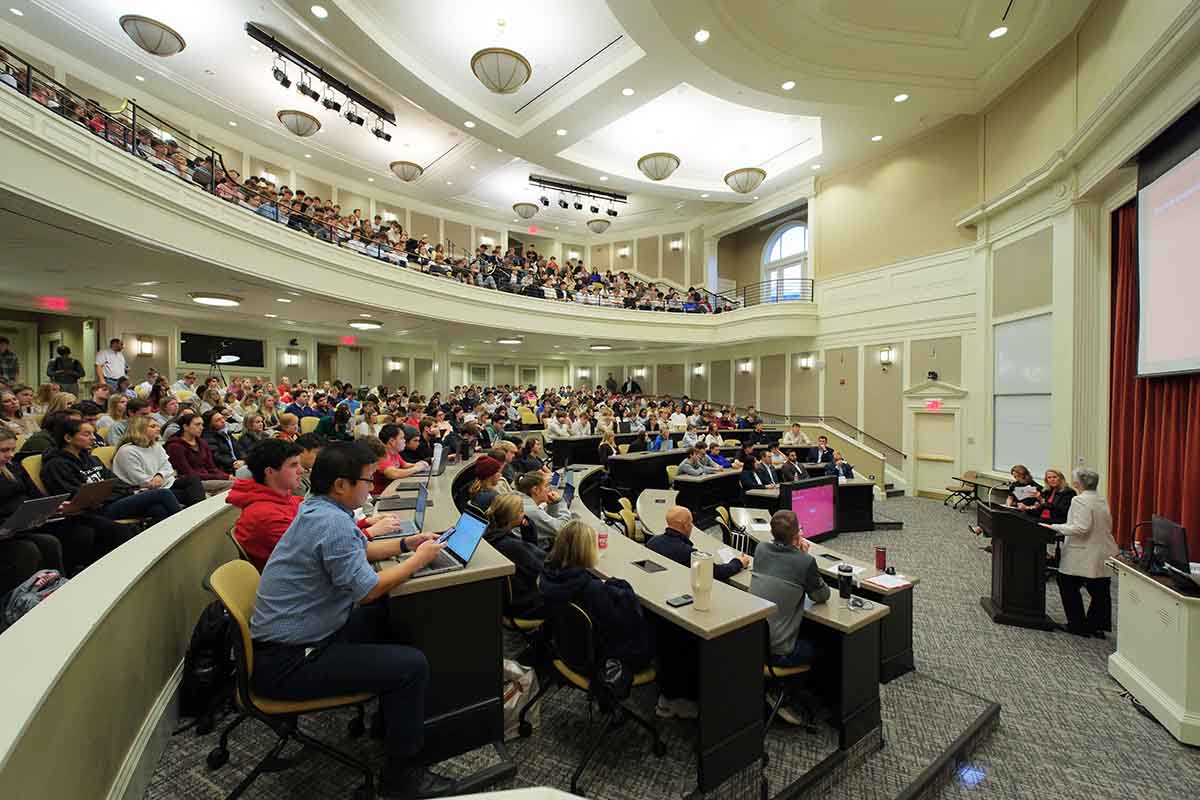 A large lecture hall filled with students attentively listening to a speaker at the front of the room.