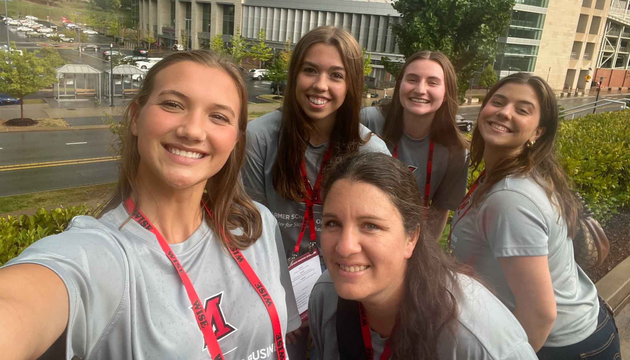 Group selfie of Murfield and students smiling, wearing matching gray t-shirts with red lanyards, outdoors at a conference.