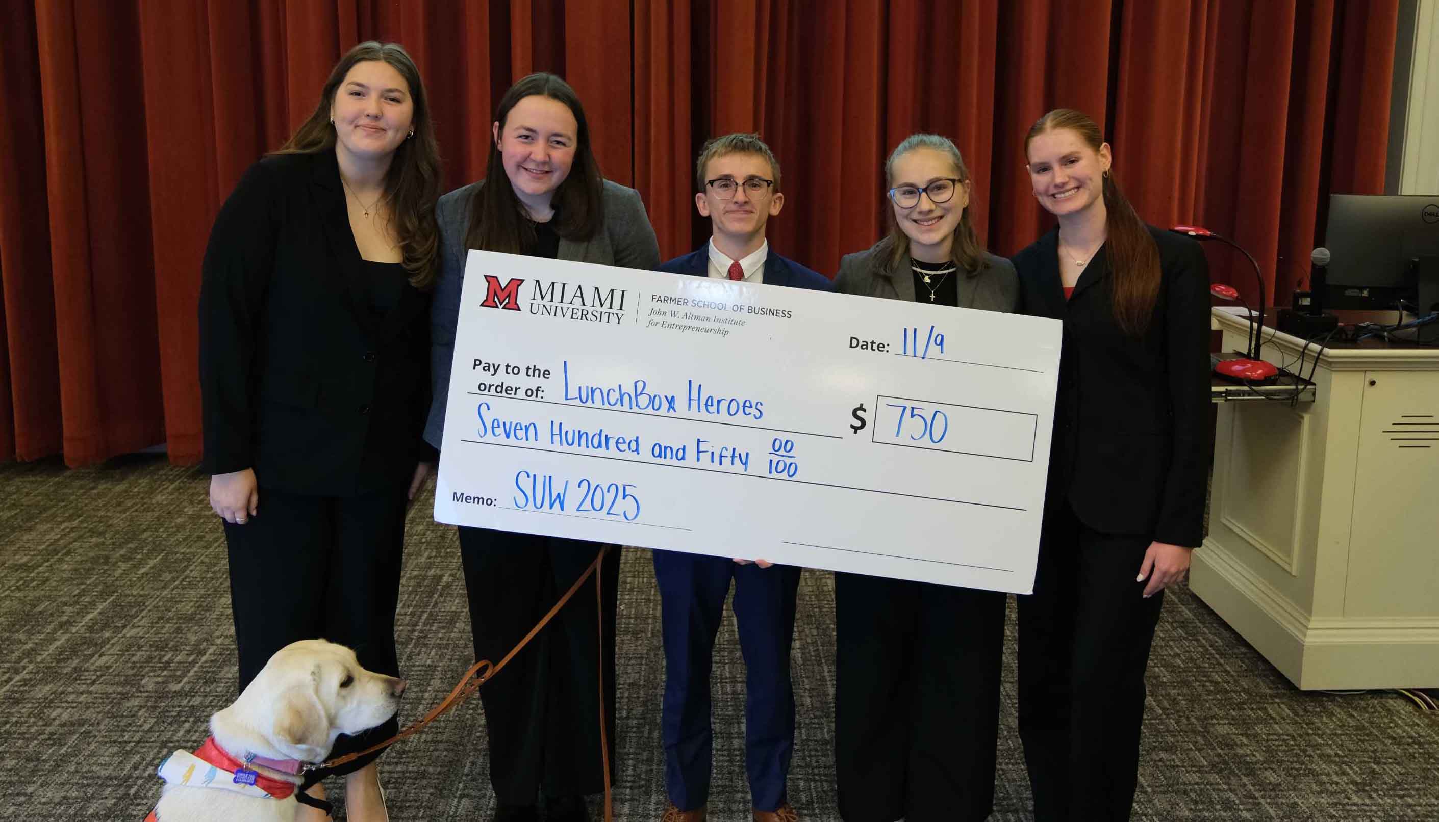 Students from Miami University holding a large check for $750, representing their idea LunchBox Heroes, with a service dog at their feet.