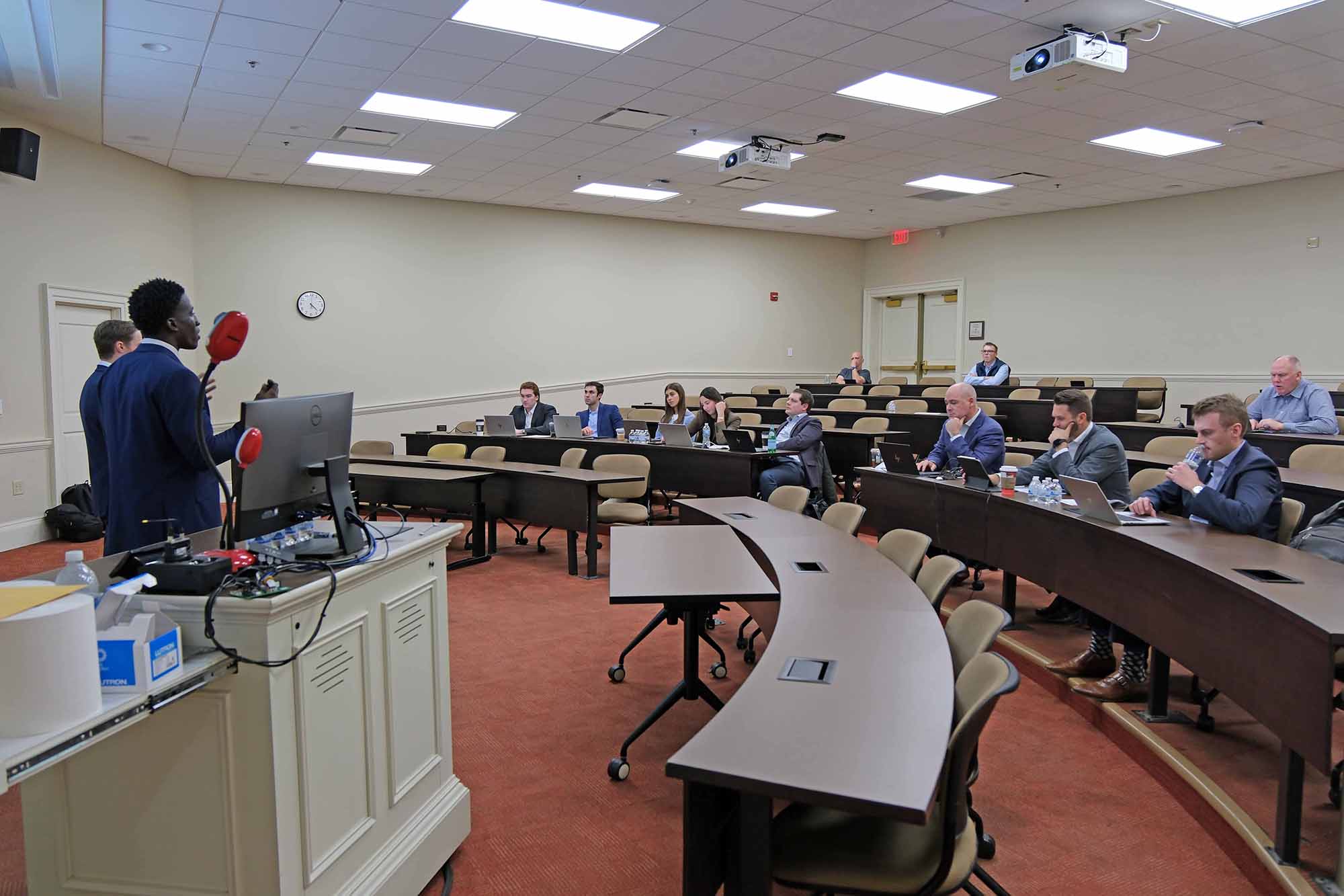 A presentation in a classroom setting with a speaker addressing an audience seated at long tables.