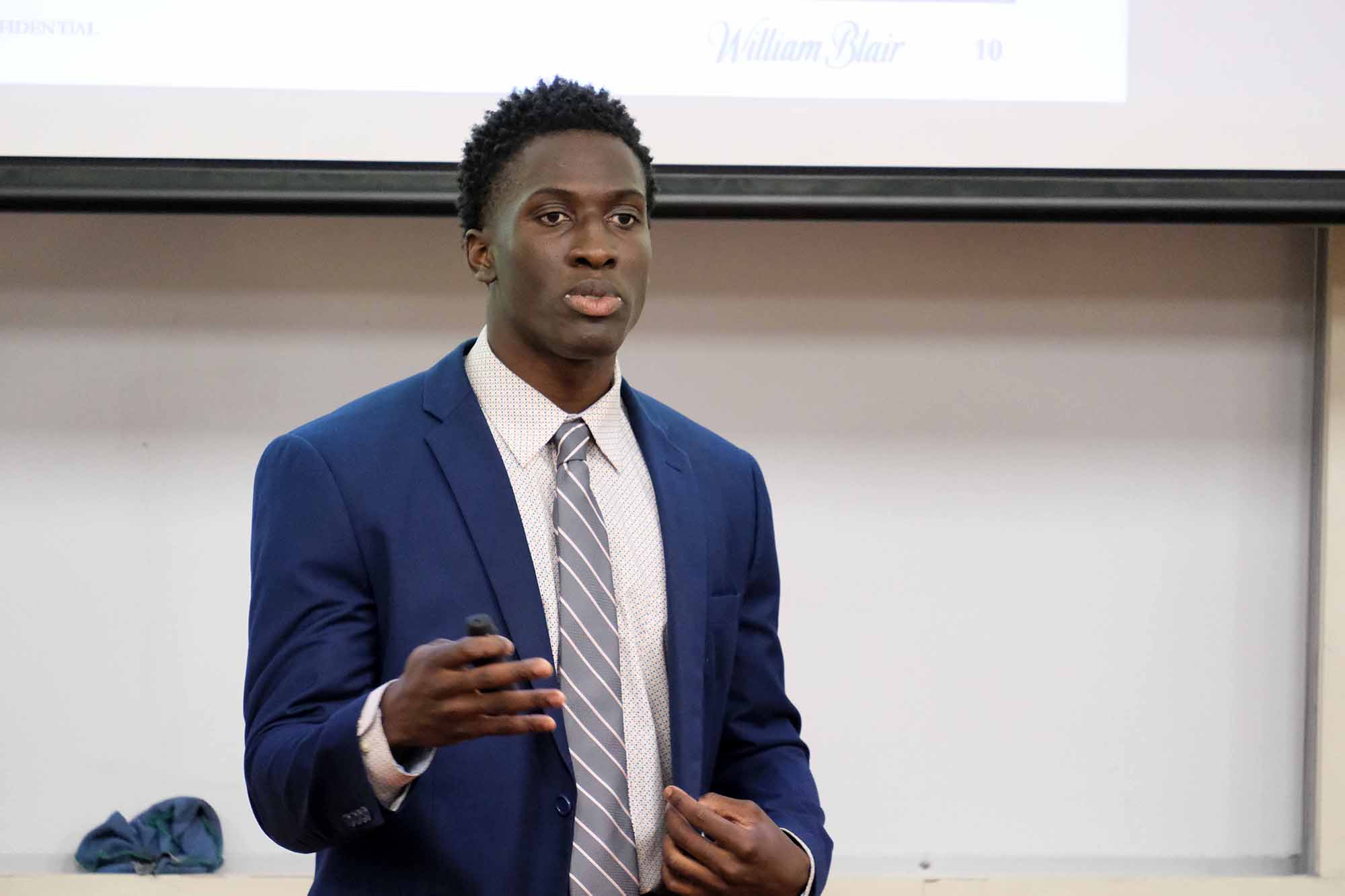 A young man in a blue suit delivers a presentation, gesturing with one hand while speaking to an audience.