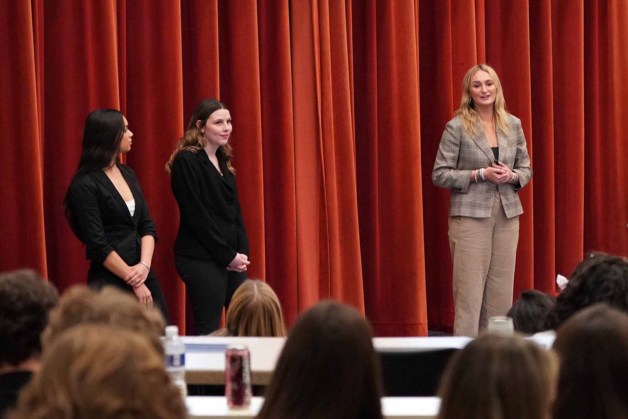 Three women present on stage in front of an audience, with a focus on one speaker addressing the crowd.