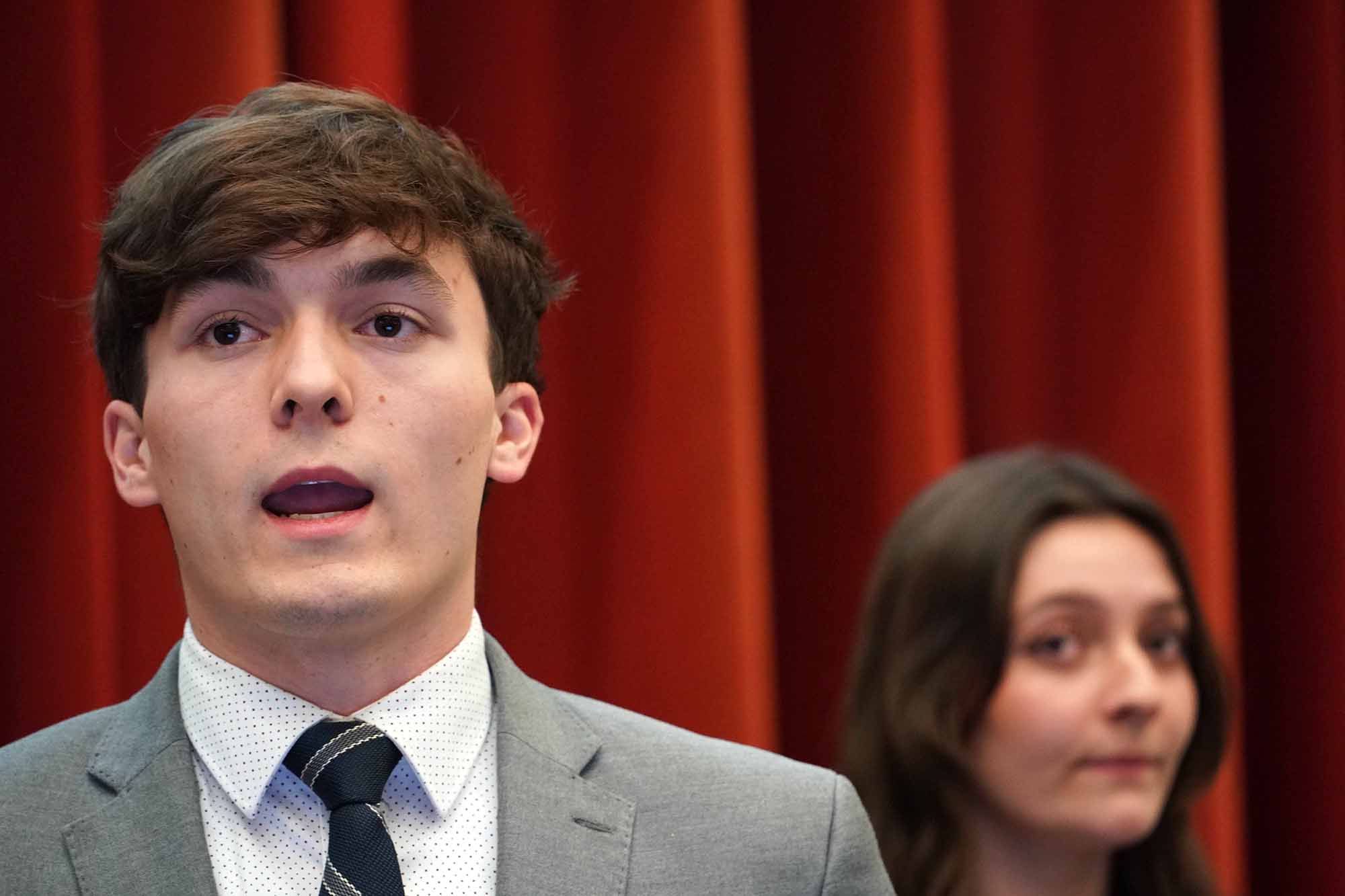 A young man in a gray suit speaks passionately at a podium, with a woman in the background observing him.