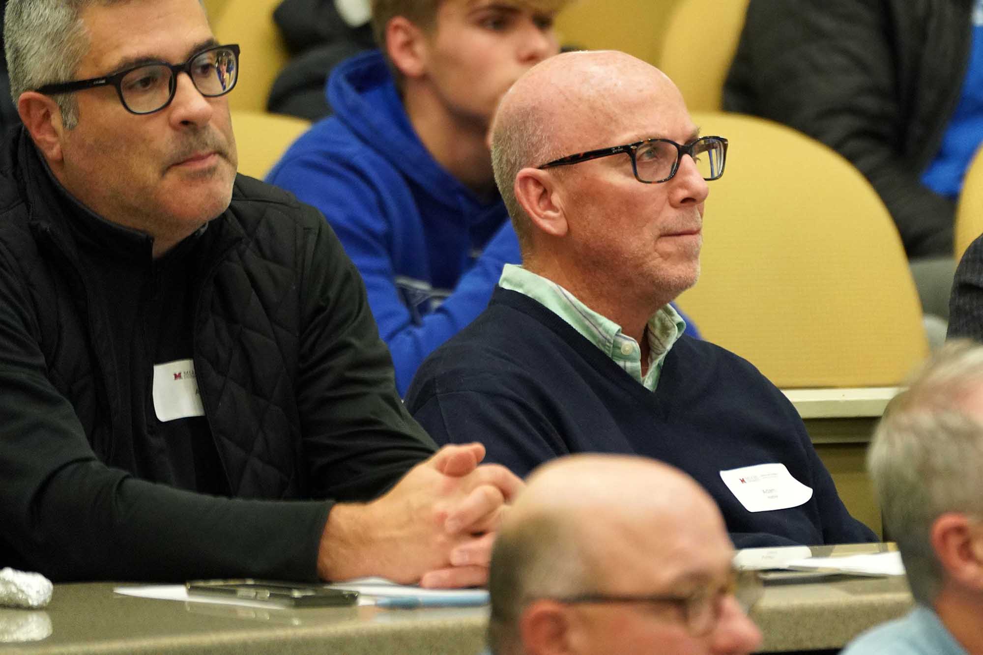 Two men are seated in an auditorium, engaged in a discussion or presentation. One man wears glasses and a dark sweater, while the other has gray hair and wears a black vest.