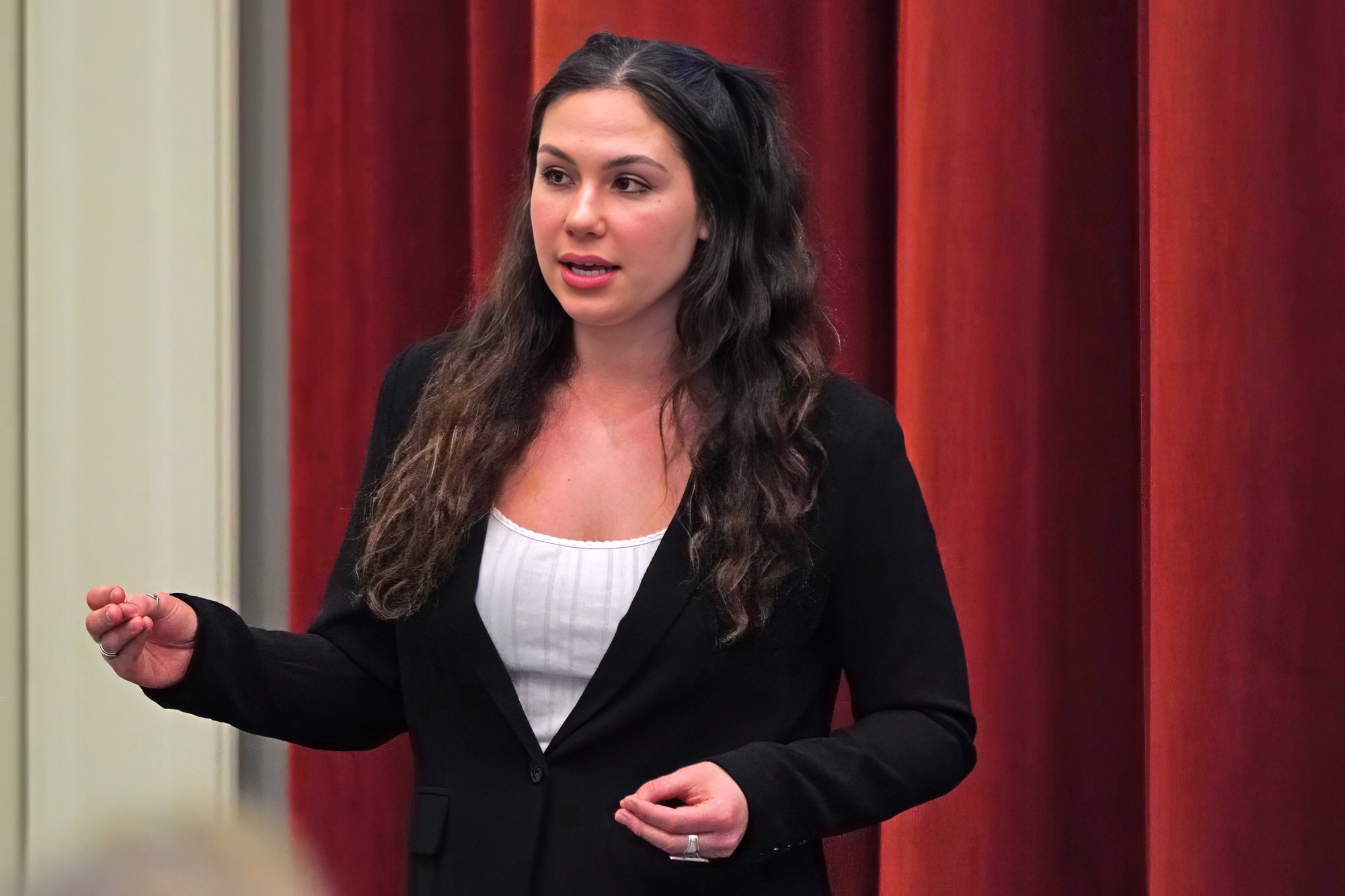 A female speaker presenting confidently in front of a red curtain.