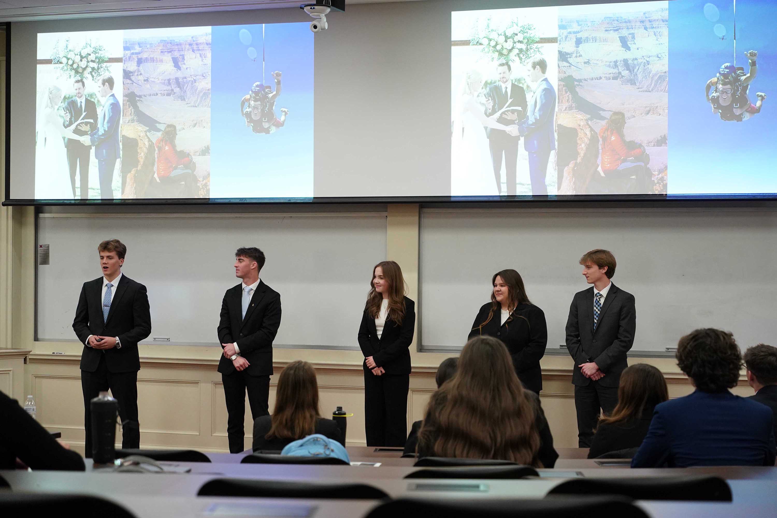 A group of five students dressed in business attire presents in front of a projector screen featuring images of outdoor activities and events.