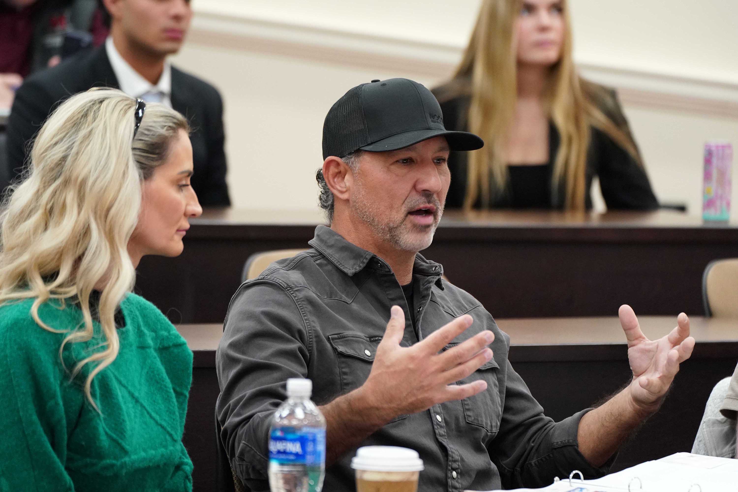 A man in a black cap gestures while speaking during a discussion in a classroom setting, with other attendees listening.