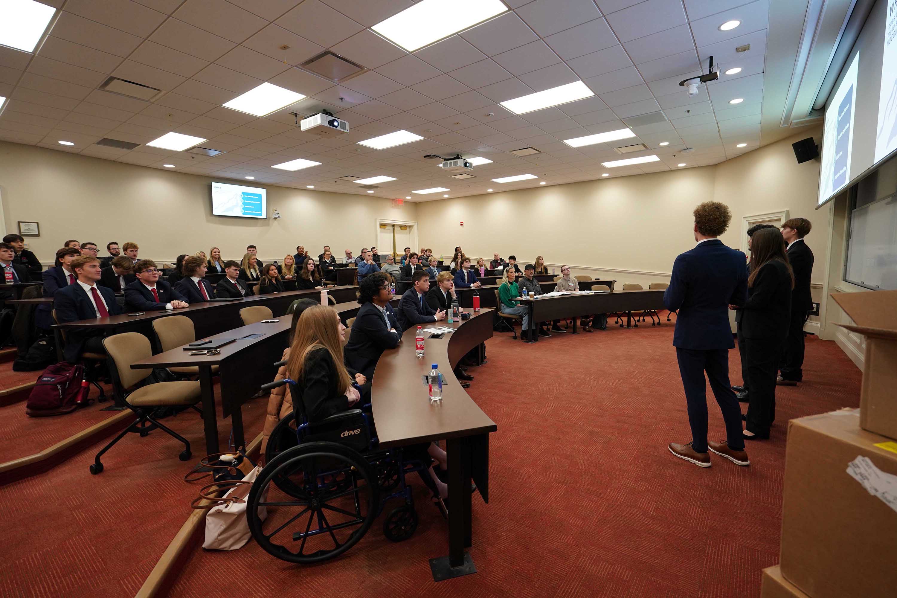 Students presenting in a classroom setting, with an audience seated in rows. The presentation includes a projector display, and some attendees are taking notes while others listen.